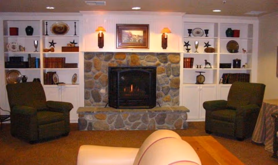 Cozy living room with a stone fireplace in the center, flanked by built-in white shelves filled with decorative items and books. Two green upholstered armchairs are positioned on either side of the fireplace, and a beige sofa is partially visible in the foreground. The room is softly lit by two wall sconces above the fireplace and recessed ceiling lights.