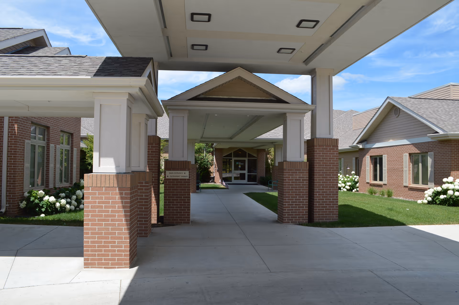 Entrance area of Parkridge Nursing & Rehab Center showing a covered drop-off zone with brick pillars and a walkway leading to the main doors. The building has brick walls and windows with white trim, surrounded by green grass and white flowering bushes under a blue sky.