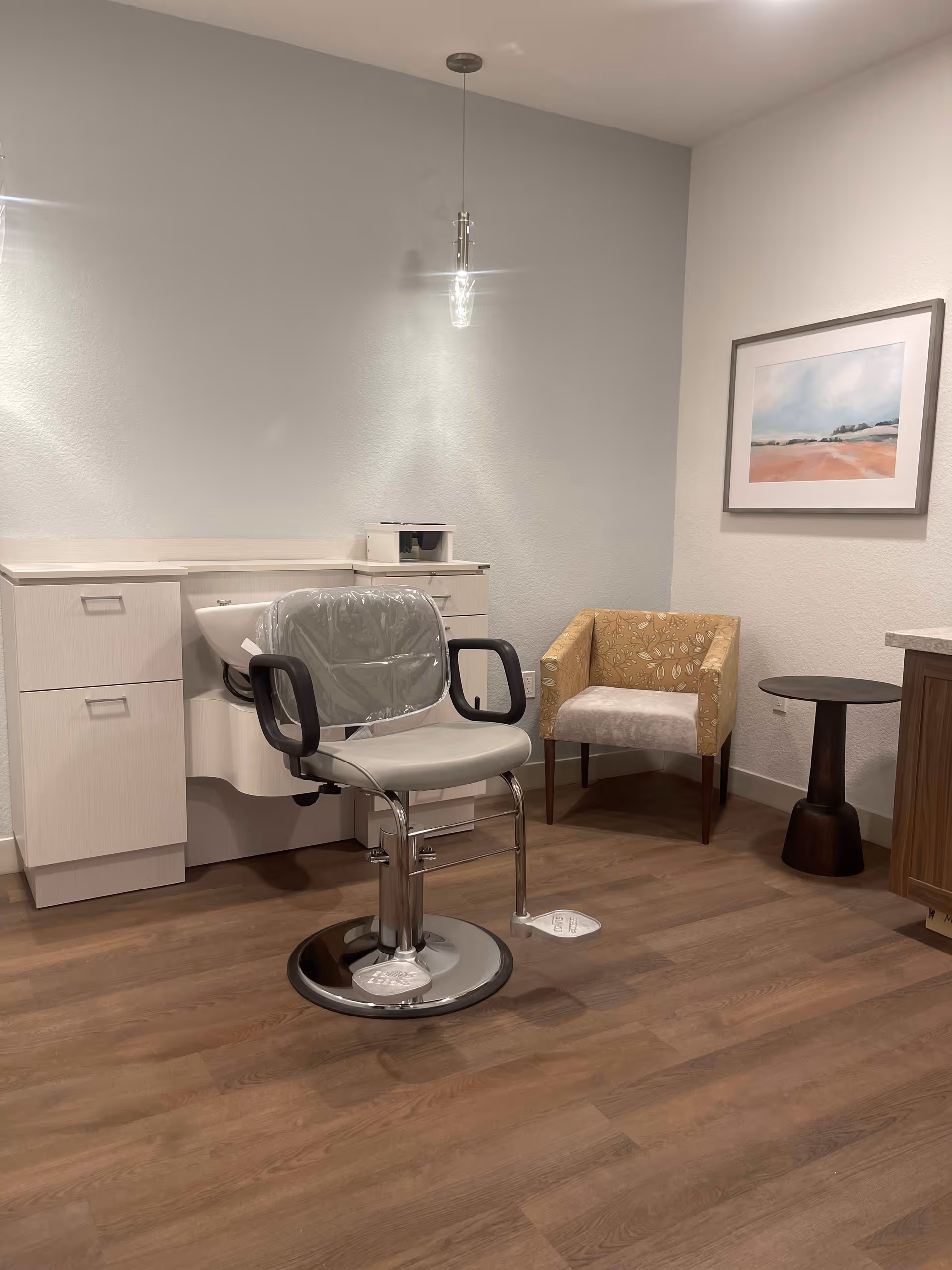 Interior room with a salon chair covered in plastic in front of a white cabinet with a sink, a yellow patterned armchair, a small dark wooden side table, and a framed landscape painting on the wall.