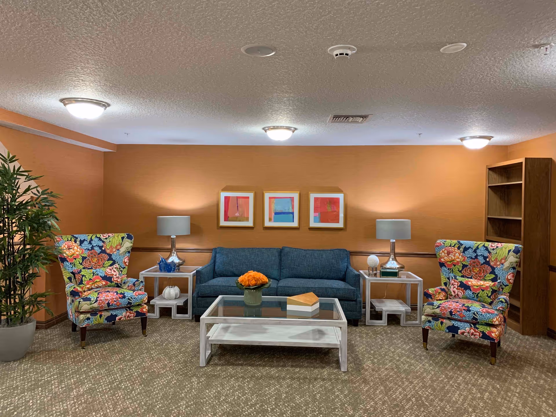 A cozy living room area with a blue sofa centered against an orange wall, flanked by two colorful floral armchairs. There are two side tables with lamps on either side of the sofa, a glass coffee table with a vase of orange flowers and a decorative box, a potted plant on the left, three abstract framed artworks on the wall, and a wooden bookshelf on the right.