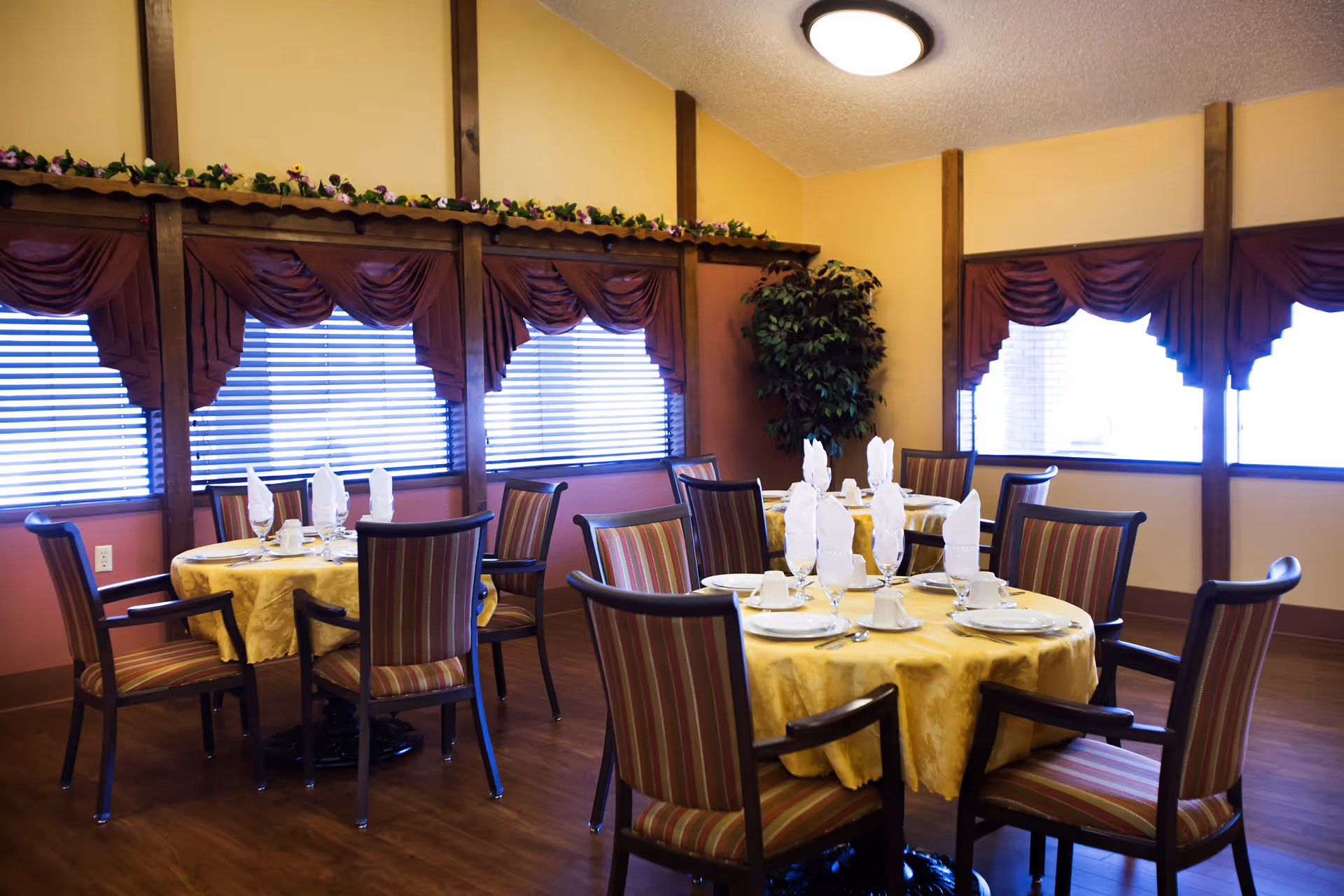 Round dining tables set with place settings and folded napkins in a warmly decorated dining room with striped chairs and curtained windows.