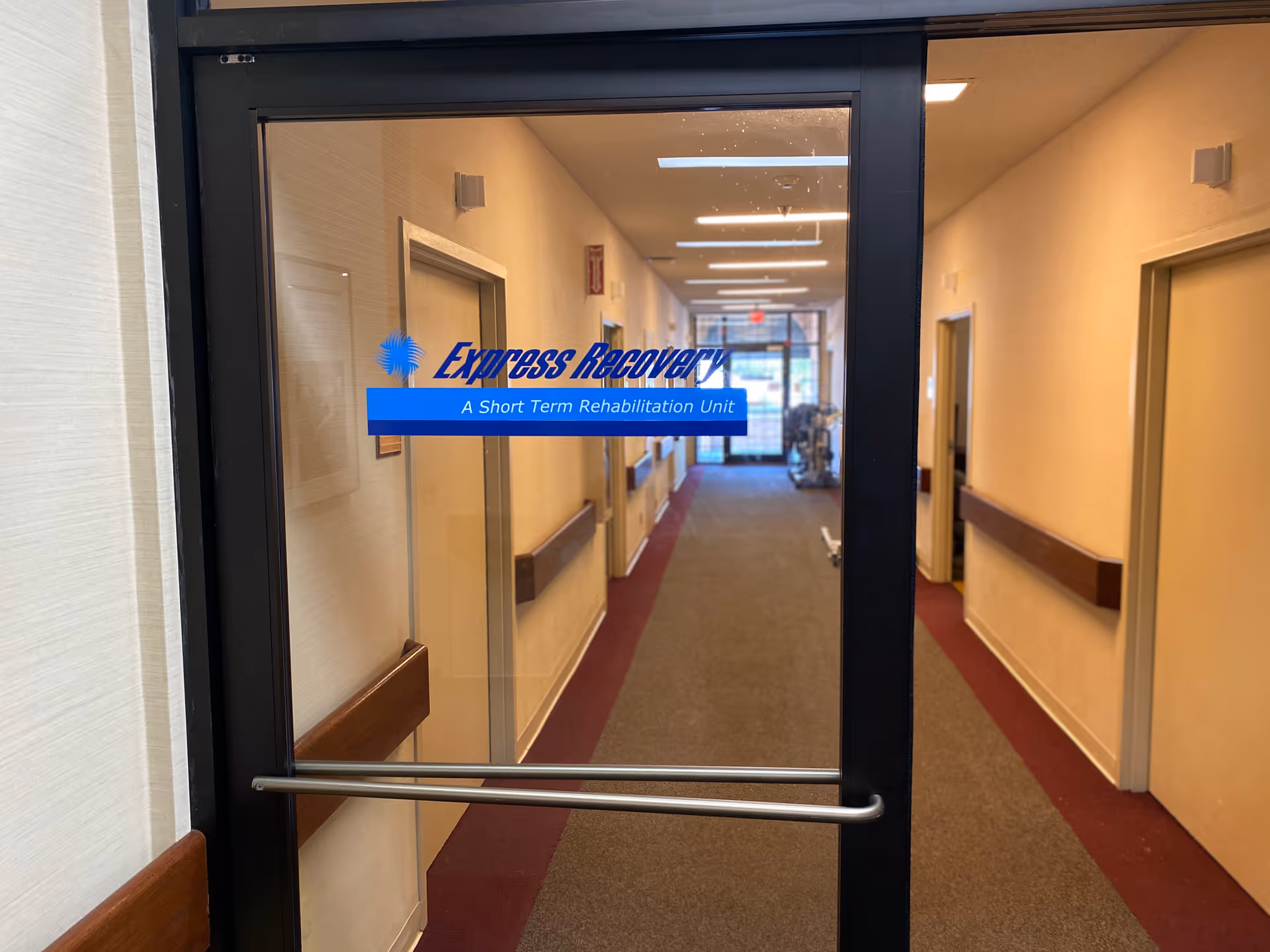 Glass door with a sign that reads 'Express Recovery A Short Term Rehabilitation Unit' leading to a hallway with beige walls, brown handrails, and several doors on each side, with a carpeted floor and ceiling lights.