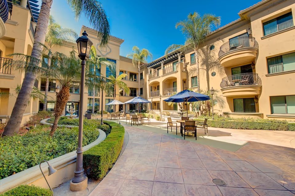 Sunlit courtyard with patio tables and blue umbrellas, palm trees, and surrounding Mediterranean-style residential building.