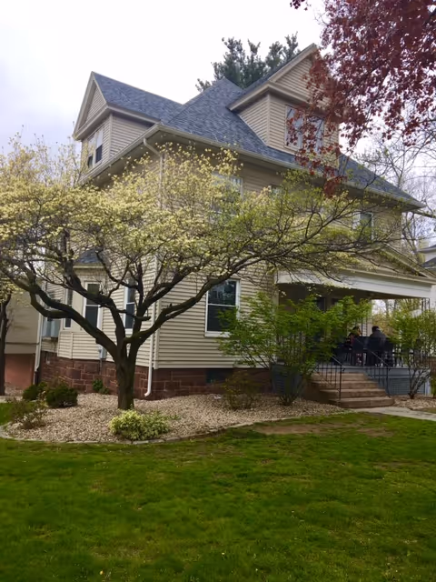 A large, beige, multi-story house with a steep roof and a front porch. The house is surrounded by green grass, small bushes, and trees with blooming flowers. There are a few people sitting on the porch. The sky is overcast.