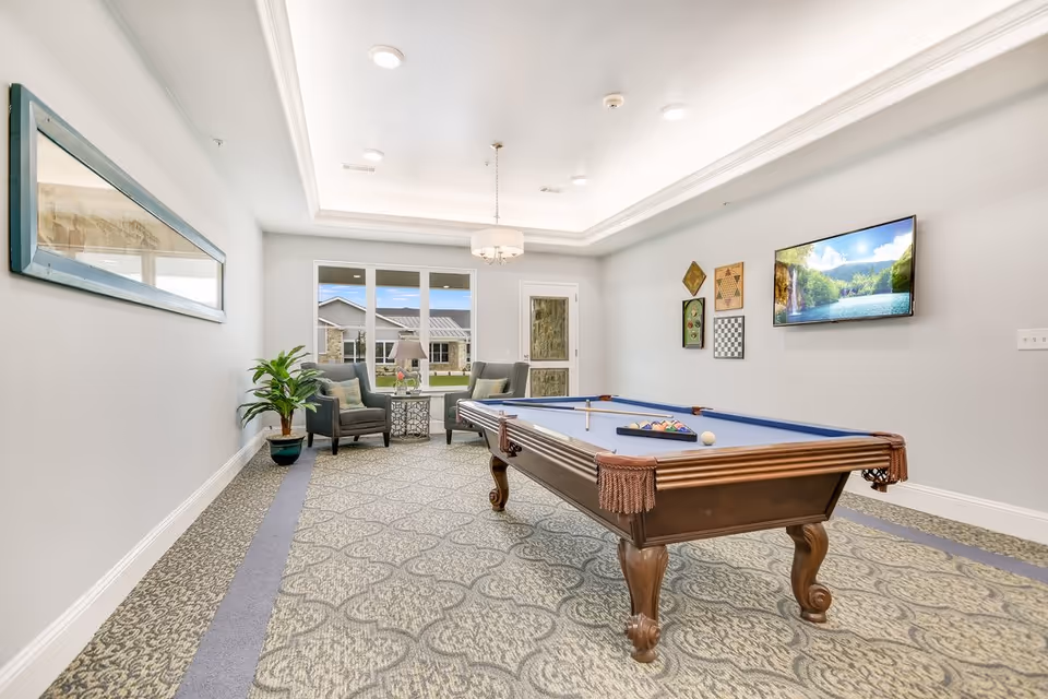 A bright and spacious game room with a pool table in the center, two armchairs with a small table and lamp near a window, a flat-screen TV mounted on the wall displaying a nature scene, and various game boards hanging on the wall. The room has patterned carpet, a large mirror on one wall, and a door leading outside.