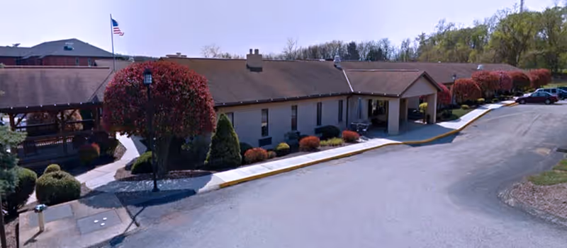 Exterior view of a single-story building with a brown roof and beige walls, surrounded by neatly trimmed bushes and trees with red foliage. A paved driveway leads to the entrance, and an American flag is visible on a flagpole near the building.