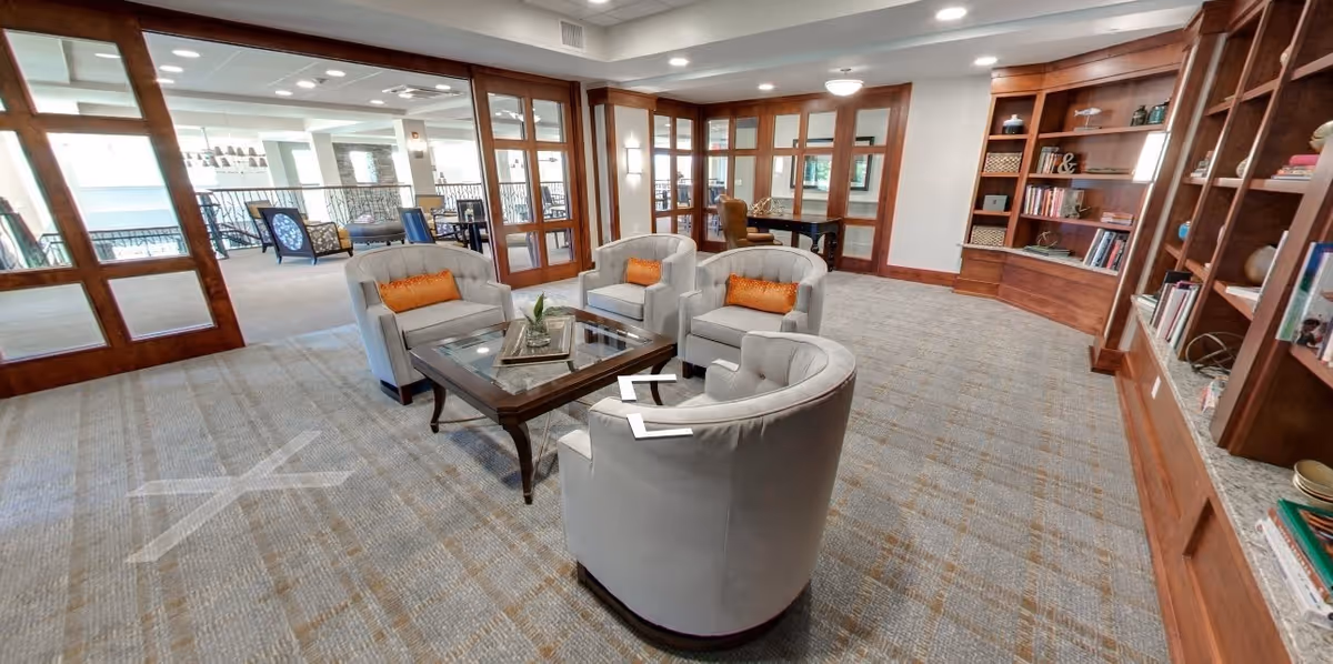 A spacious and well-lit lounge area with four light gray armchairs arranged around a glass-top coffee table. The room features wood-framed glass doors, built-in wooden bookshelves filled with books and decorative items, and a carpeted floor. In the background, there is an adjacent seating area with additional chairs and tables.