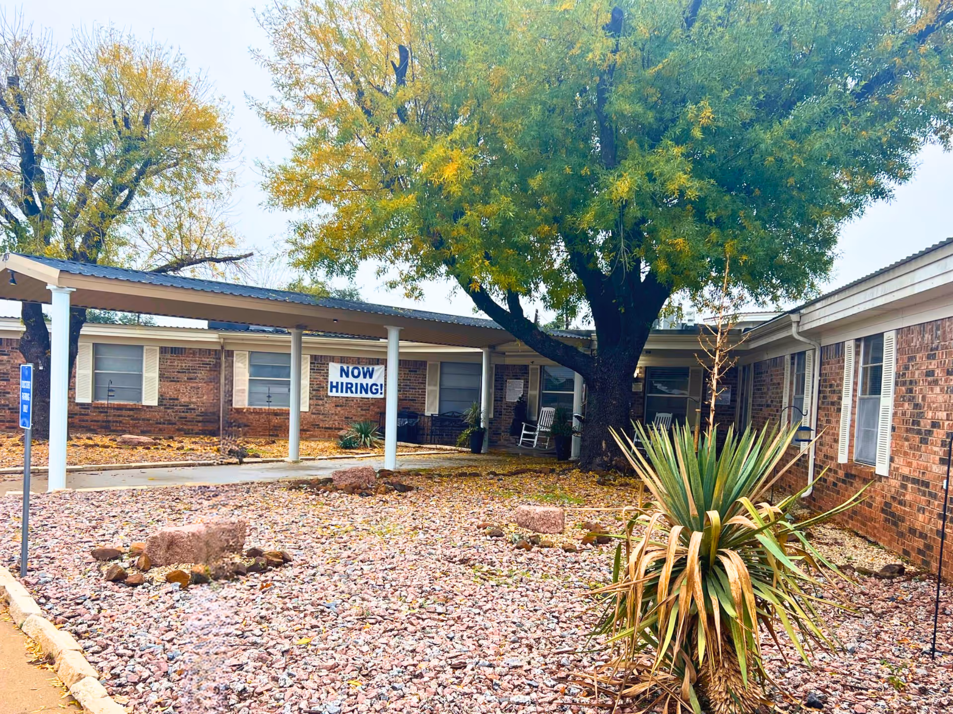 Front exterior of a single-story brick nursing facility with a covered entrance, large tree, rock landscaping, and a 'NOW HIRING!' sign.