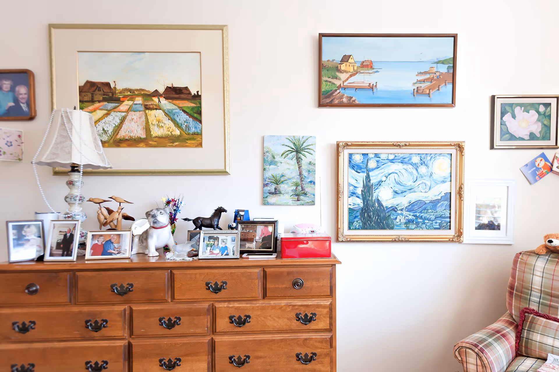 Wooden dresser topped with framed photos, a lamp and decorative figurines beneath a wall of framed paintings, beside a plaid armchair.