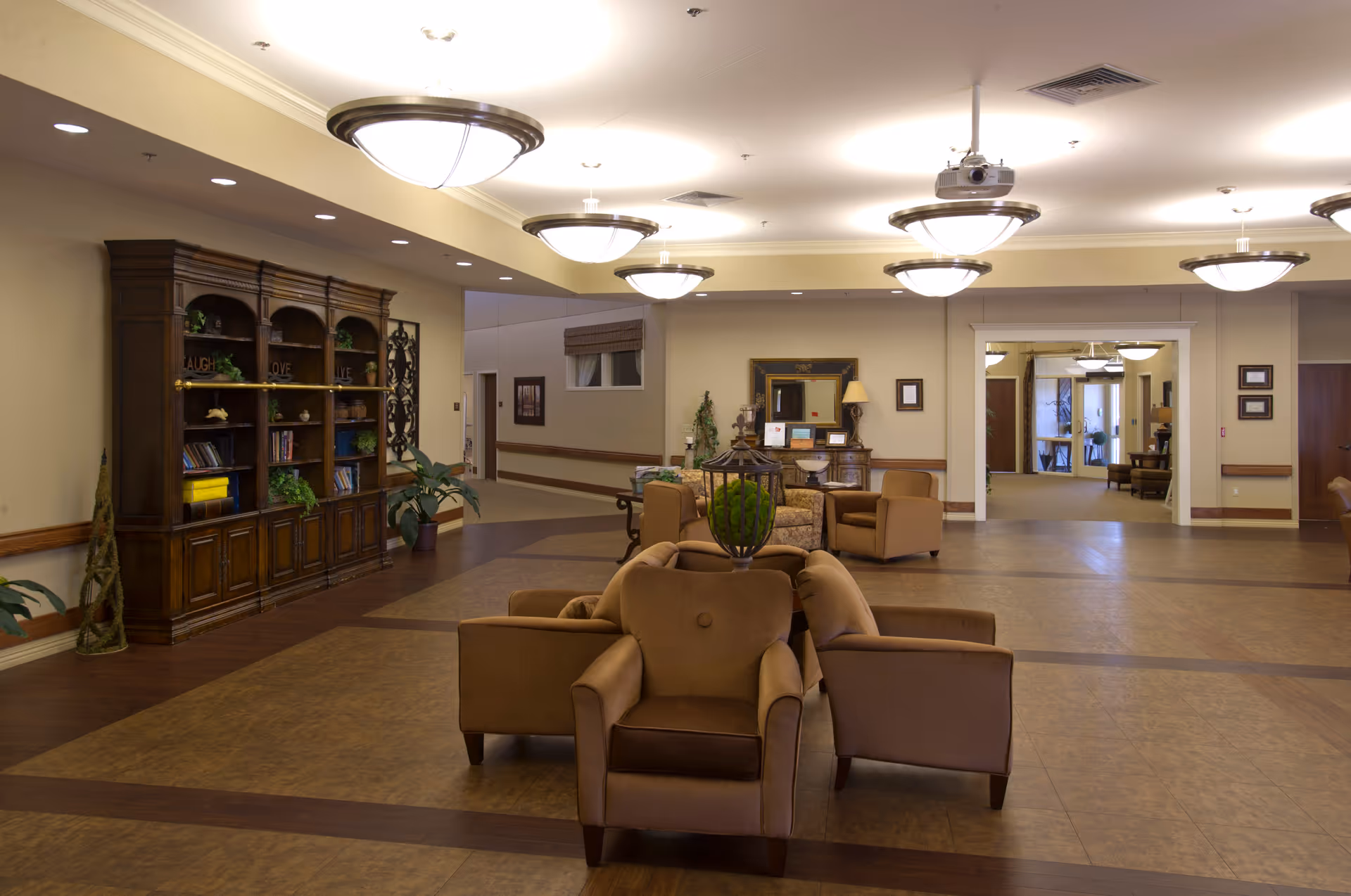 A spacious and well-lit common area in a senior living facility featuring comfortable brown armchairs arranged around a central decorative piece. The room has a large wooden bookshelf filled with books and decorative items on the left side, and multiple ceiling lights providing warm illumination. There is a hallway and additional seating visible in the background.