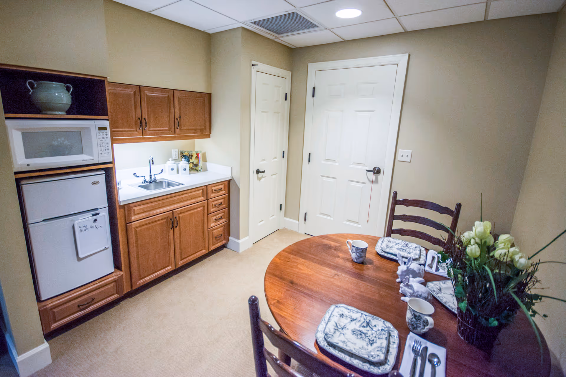 Small kitchenette with wooden cabinets, a sink, microwave and mini fridge beside a round dining table set with plates, cups and a floral centerpiece.