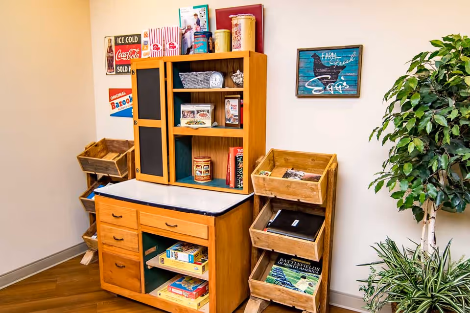 A cozy corner of an interior room featuring a wooden cabinet with shelves and drawers filled with books, games, and decorative items. On the wall above the cabinet are vintage-style signs, including one advertising Coca-Cola and another with a chicken and the words 'Farm Fresh Eggs.' To the right, there is a tall green leafy plant and a smaller plant on the floor. The floor is wooden, and the walls are painted a light color.