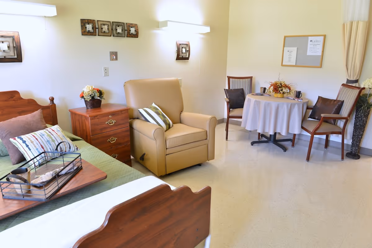 A cozy room with a wooden bed featuring pillows and a tray, a wooden nightstand with a flower pot, a beige recliner chair with a striped pillow, and a small round table covered with a tablecloth surrounded by three wooden chairs with cushions. The walls are decorated with framed art and a bulletin board.