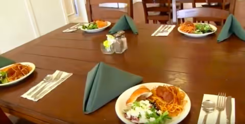 A dining table set with plates of food including salad, bread, and pasta. Each place setting has a folded green napkin, fork, knife, and spoon. Salt and pepper shakers are placed in the center of the table.