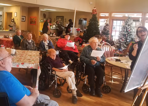 A group of elderly people, some in wheelchairs, seated in a common room decorated with Christmas trees and holiday decorations. A woman stands at a whiteboard, engaging with the group, who are holding papers. The room has wooden floors and large windows letting in natural light.