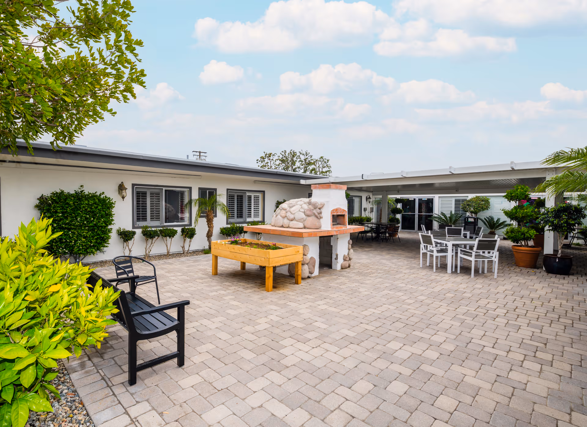 Outdoor courtyard area at Anaheim Healthcare Center featuring a stone fireplace with a built-in oven, a wooden planter box, black and white chairs and tables, potted plants, and a paved stone floor under a partly cloudy sky.