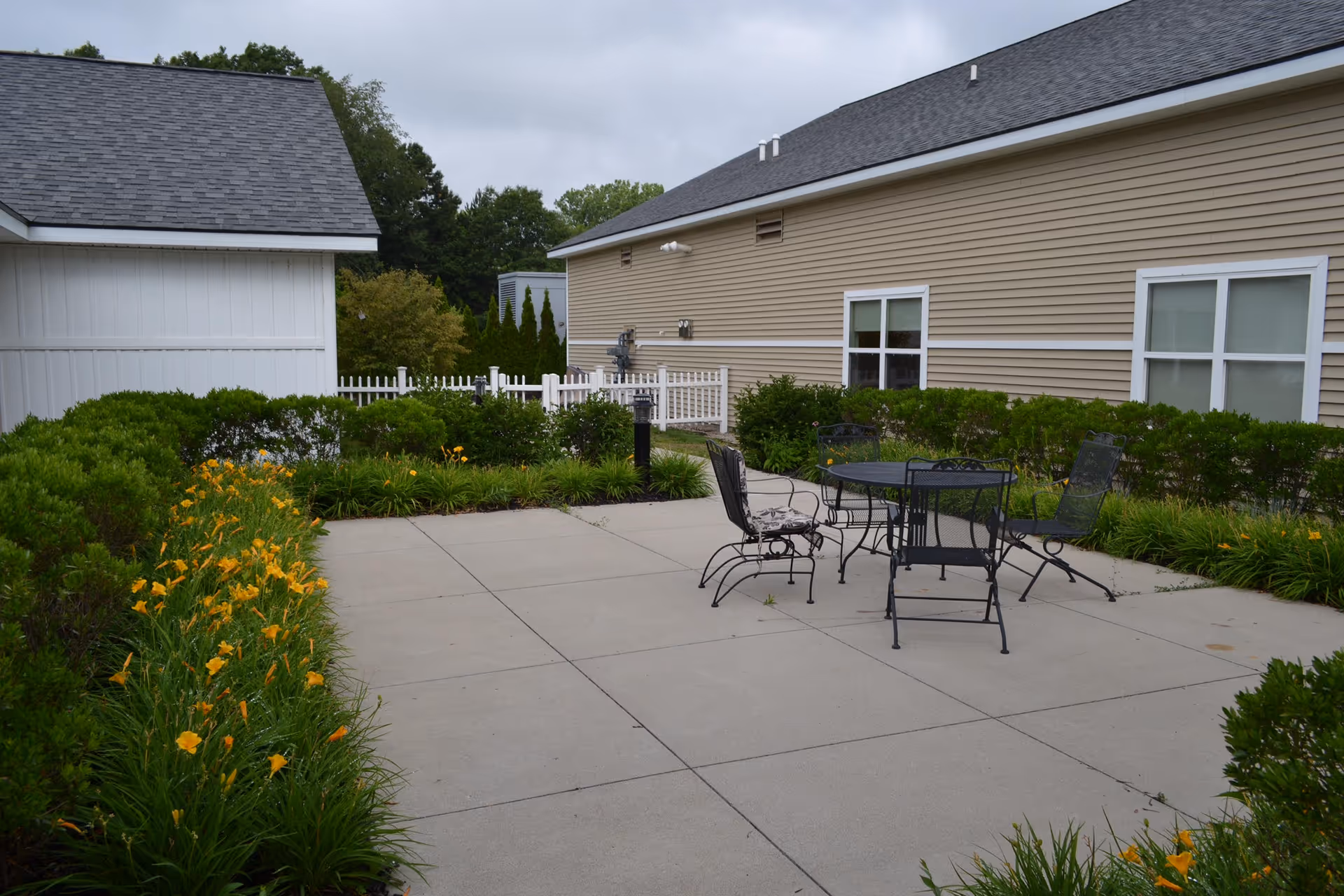 Outdoor patio area at Medilodge of Montrose featuring a concrete floor with a black metal table and four chairs. The patio is surrounded by green bushes and yellow flowers, with beige siding buildings and white fencing in the background under a cloudy sky.