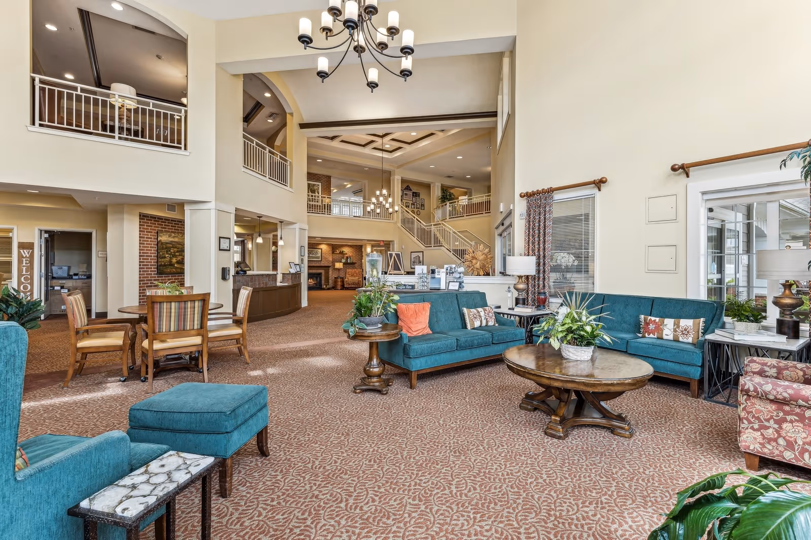 Spacious senior living facility common area with high ceilings, teal sofas, patterned carpet, wooden coffee tables, and indoor plants. There is a staircase leading to an upper level with railings, a reception desk, and a seating area with a round table and chairs. Large windows and warm lighting create a welcoming atmosphere.
