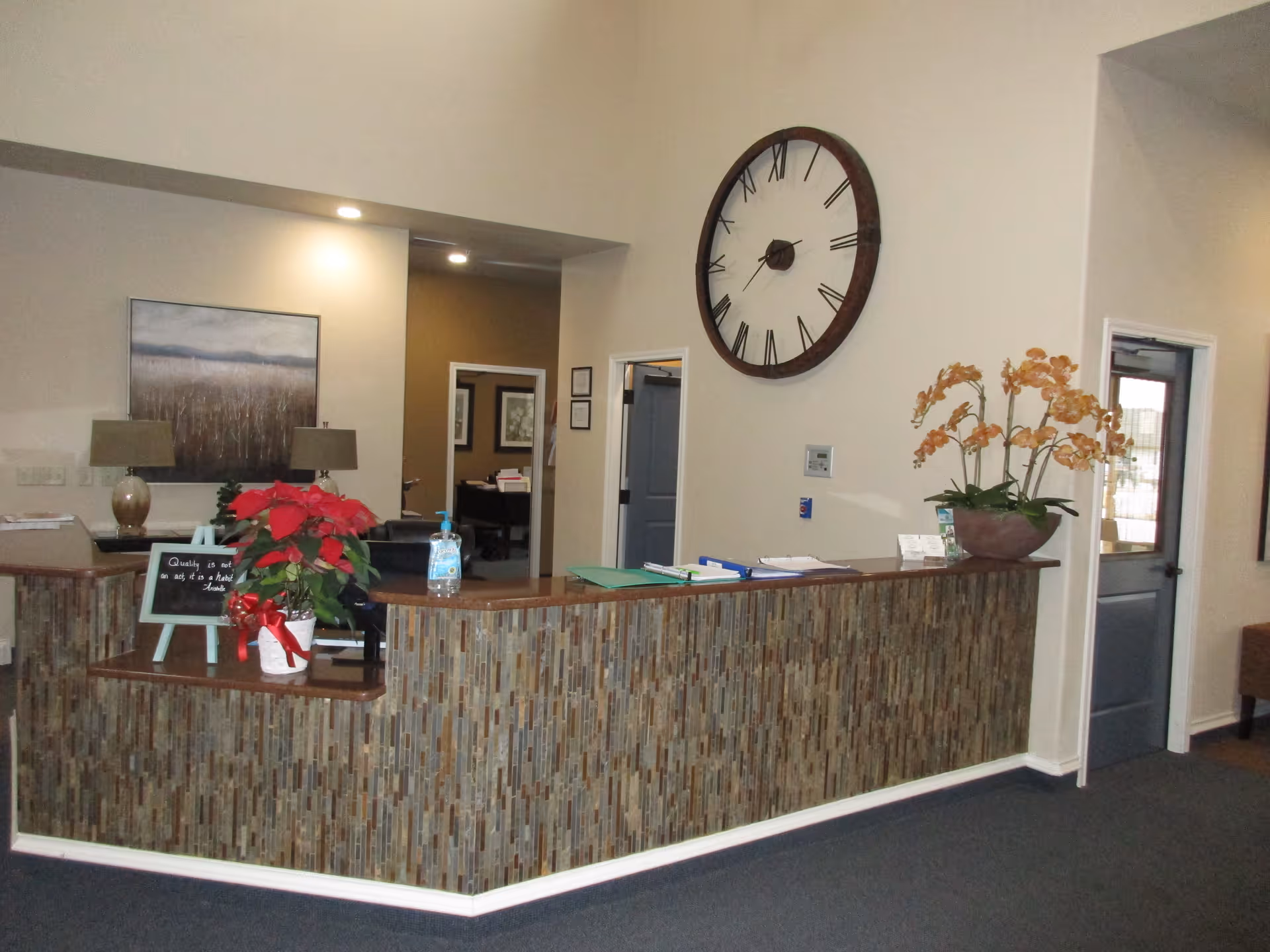 Reception desk area in an assisted living facility with a large wall clock above, a poinsettia plant with a red bow, a hand sanitizer bottle, and a potted orchid on the counter. There are two table lamps and a framed landscape painting on the wall behind the desk. Two doorways lead to other rooms.