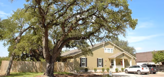 Single-story beige building with white trim and a small front porch, surrounded by a wooden fence and large leafy tree in the front yard. Several cars are parked in the driveway under a clear blue sky.