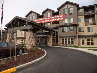 Exterior view of a multi-story senior living facility with a covered entrance. The building has a combination of stone and siding on the facade, with multiple windows and balconies. A red banner hangs above the entrance displaying the words 'Senior Living' and a phone number. There is a driveway leading up to the entrance and a parked car visible on the left side.