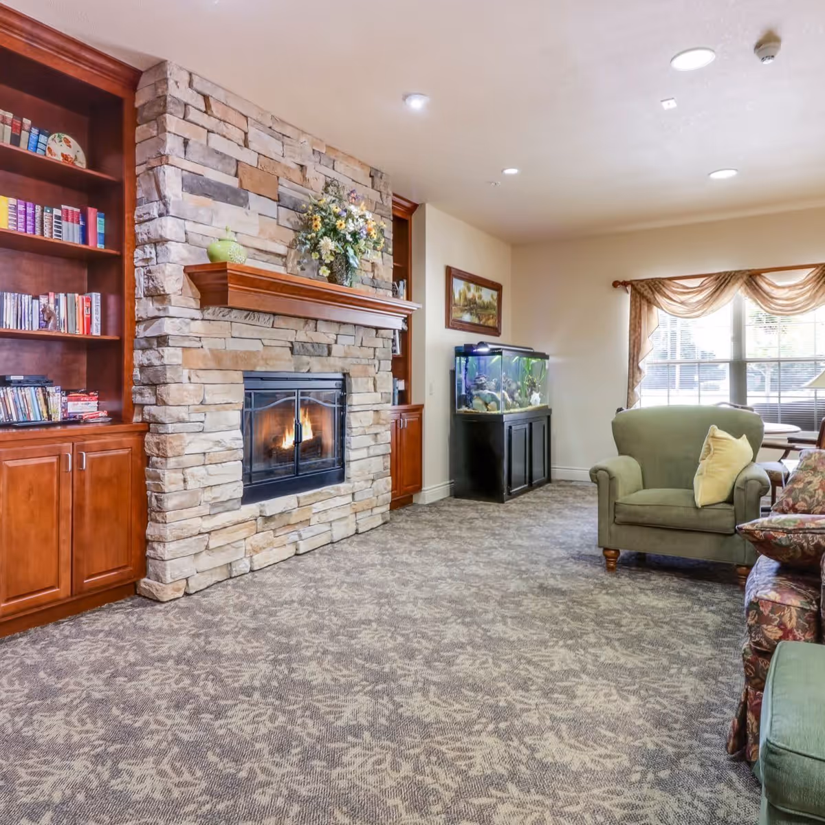 A cozy living room area featuring a stone fireplace with a wooden mantel decorated with a flower arrangement. To the left of the fireplace is a built-in wooden bookshelf filled with books and decorative items. On the right side of the room, there is a large fish tank on a black stand, a green armchair with a yellow pillow, and a window with sheer curtains allowing natural light to enter. The floor is carpeted with a patterned design.
