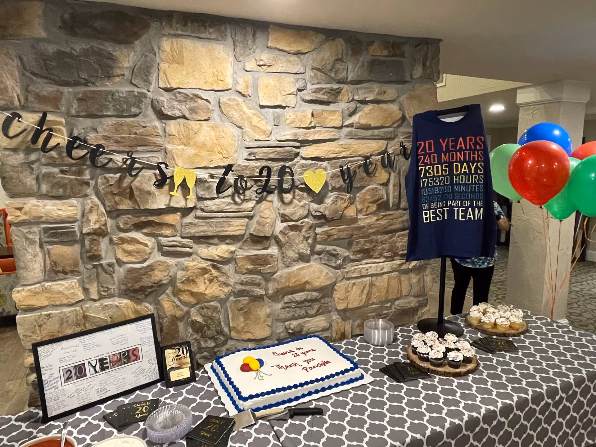 A decorated celebration table with cake, cupcakes, balloons and a banner hung on a stone wall.