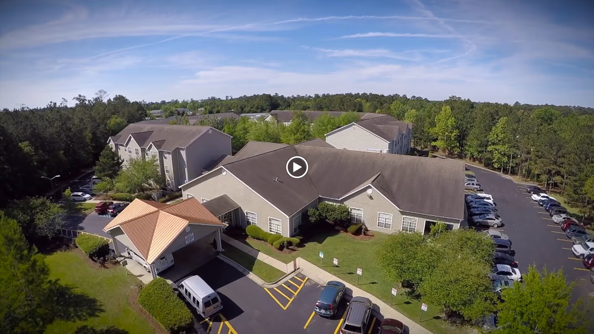 Aerial view of The Windsor Senior Living Community showing multiple connected buildings with gray roofs surrounded by trees and greenery. There is a parking lot with several cars parked, and a covered entrance with a tan roof near the front of the main building. The sky is clear with some wispy clouds.