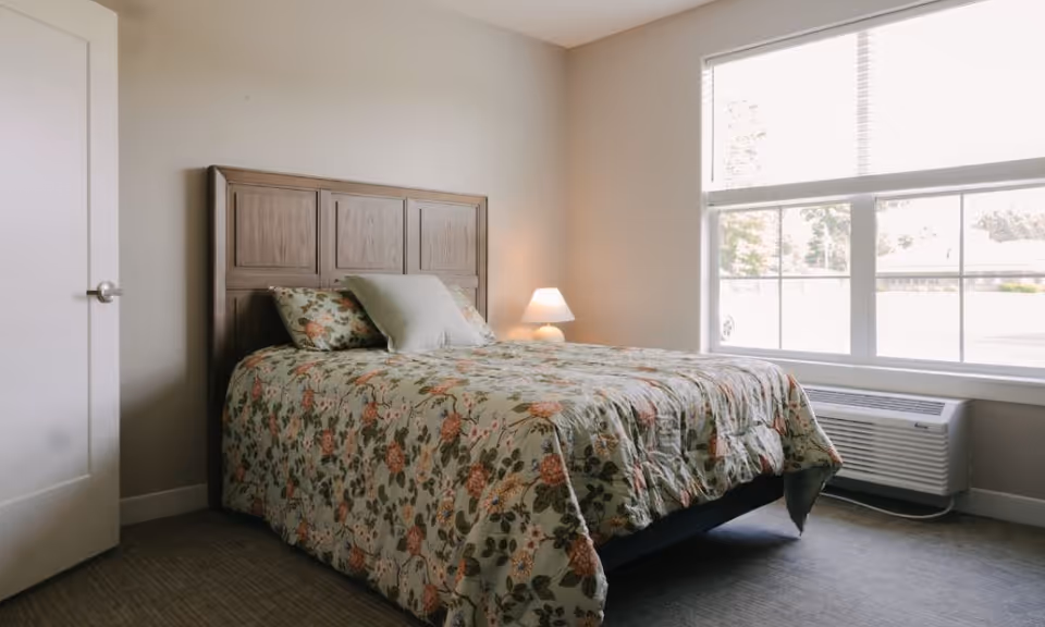 A bedroom with a wooden headboard bed covered with a floral patterned comforter and pillows. There is a small lamp on a nightstand next to the bed, a large window with blinds letting in natural light, and a white door on the left side of the room.