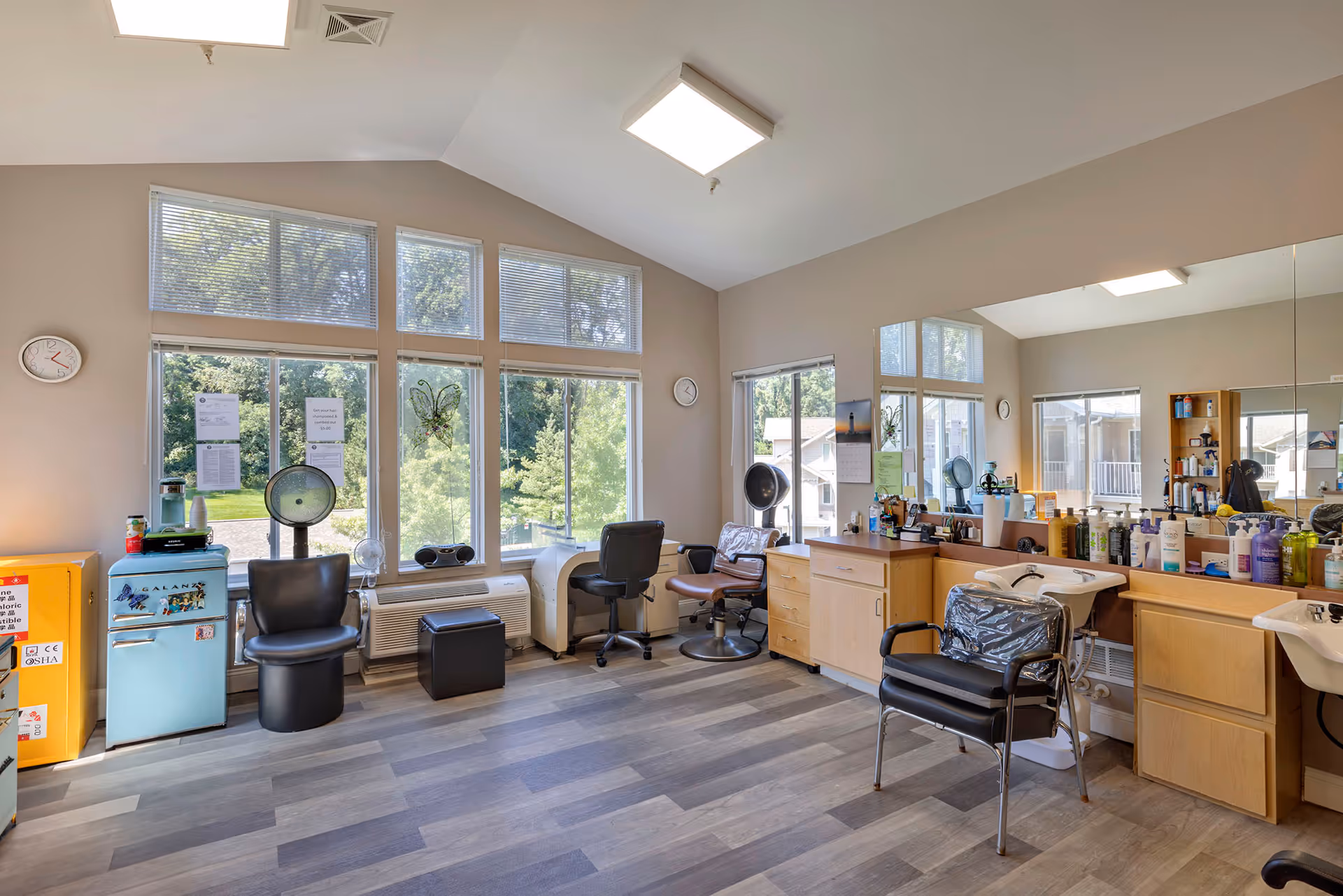 Interior of a hair salon room with large windows letting in natural light. The room features salon chairs, hair dryers, a small blue refrigerator, cabinets with various hair care products, and a large mirror along one wall. The floor is wood-patterned, and there are clocks on the walls.