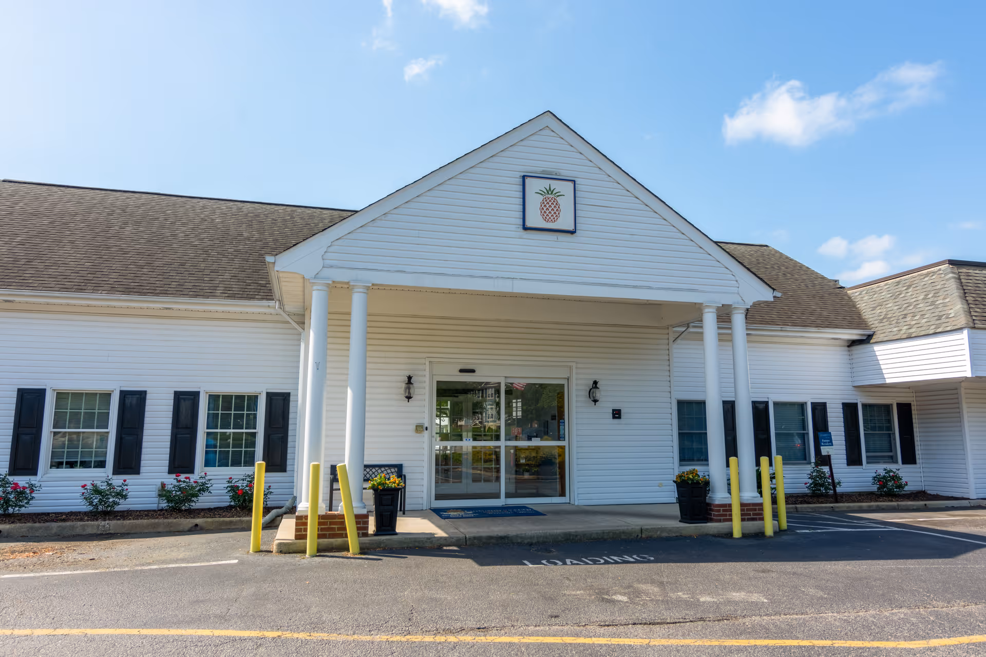 Front entrance of a white memory care building with columns, sliding glass doors, planters, and a pineapple emblem above the portico.