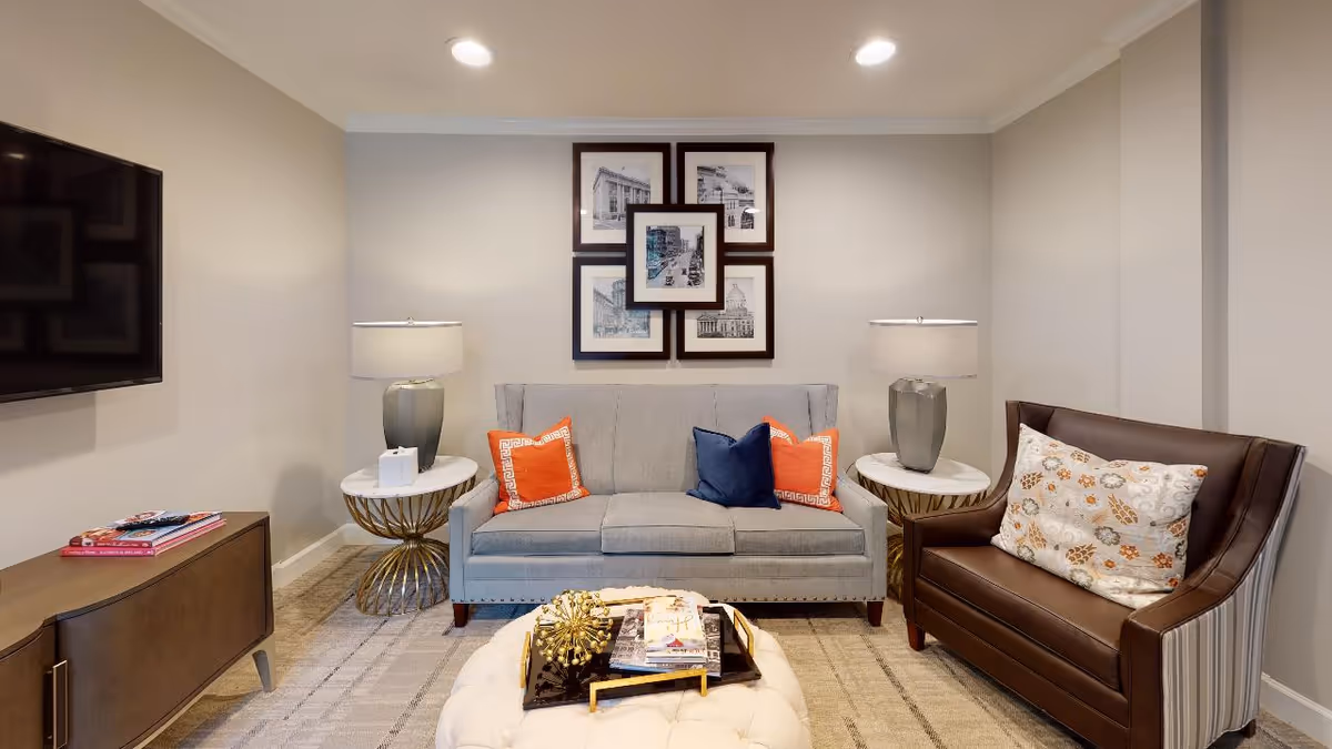 A cozy living room with a gray sofa adorned with two orange and one blue throw pillows, flanked by two round side tables each with a lamp. To the right is a brown leather armchair with a patterned cushion. A tufted white ottoman with books and decorative items sits in front of the sofa. On the wall above the sofa are five framed black and white architectural sketches arranged in a grid. A flat-screen TV is mounted on the left wall above a wooden cabinet with magazines on top. The room has beige walls and recessed ceiling lights.