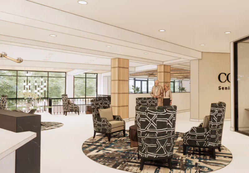 Bright senior living lobby with patterned armchairs arranged on circular rugs, large windows, wooden columns, and two people near the reception area.