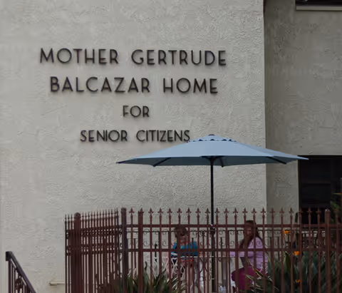 Exterior wall of the Mother Gertrude Balcazar Home for Senior Citizens with its name displayed above a fenced patio area with an umbrella and two seated people.