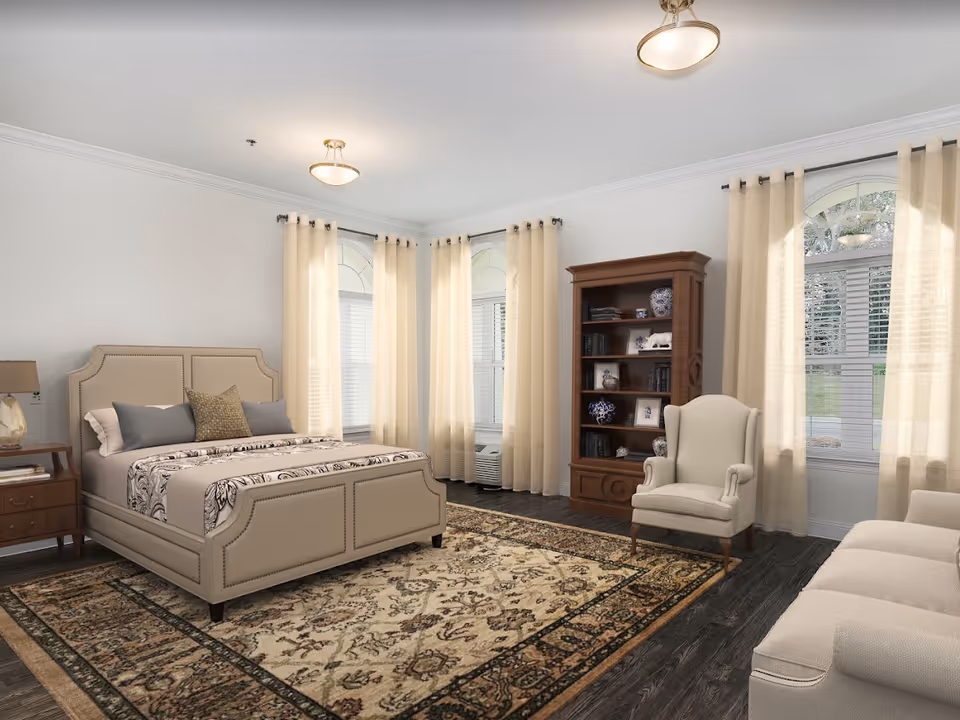 A spacious bedroom with a beige upholstered bed, patterned bedding, and multiple pillows. The room features three large windows with sheer beige curtains, a wooden bookshelf filled with decorative items and books, a beige armchair, a beige sofa, a wooden nightstand with a lamp, and a large patterned area rug on dark wood flooring.