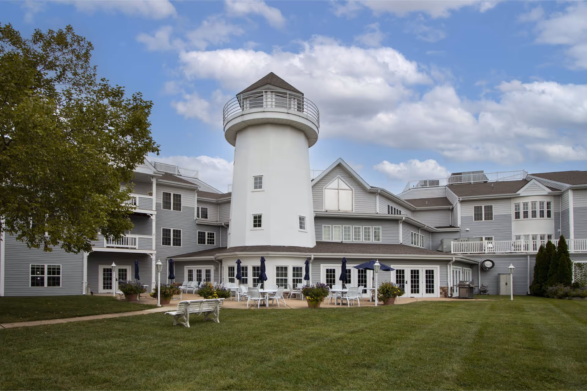 Exterior view of Cape Albeon Independent Living facility featuring a large white lighthouse structure in the center, surrounded by a multi-story gray building with white trim. There is a patio area with tables, chairs, and umbrellas, and a well-maintained lawn with a bench in the foreground under a partly cloudy sky.