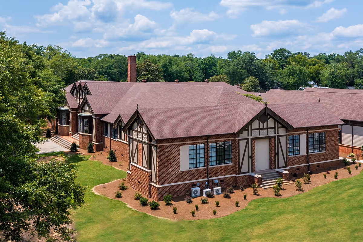 Exterior view of a large brick building with Tudor-style architectural details, surrounded by green grass and trees under a partly cloudy sky.