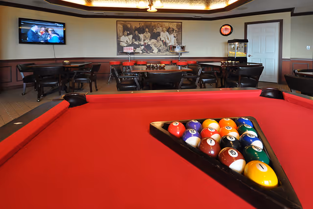 Interior view of a recreational room featuring a red pool table with a full rack of billiard balls in the foreground. In the background, there are several tables and chairs, a wall-mounted TV showing a scene with people, a vintage-style photograph on the wall, a popcorn machine, and a door.