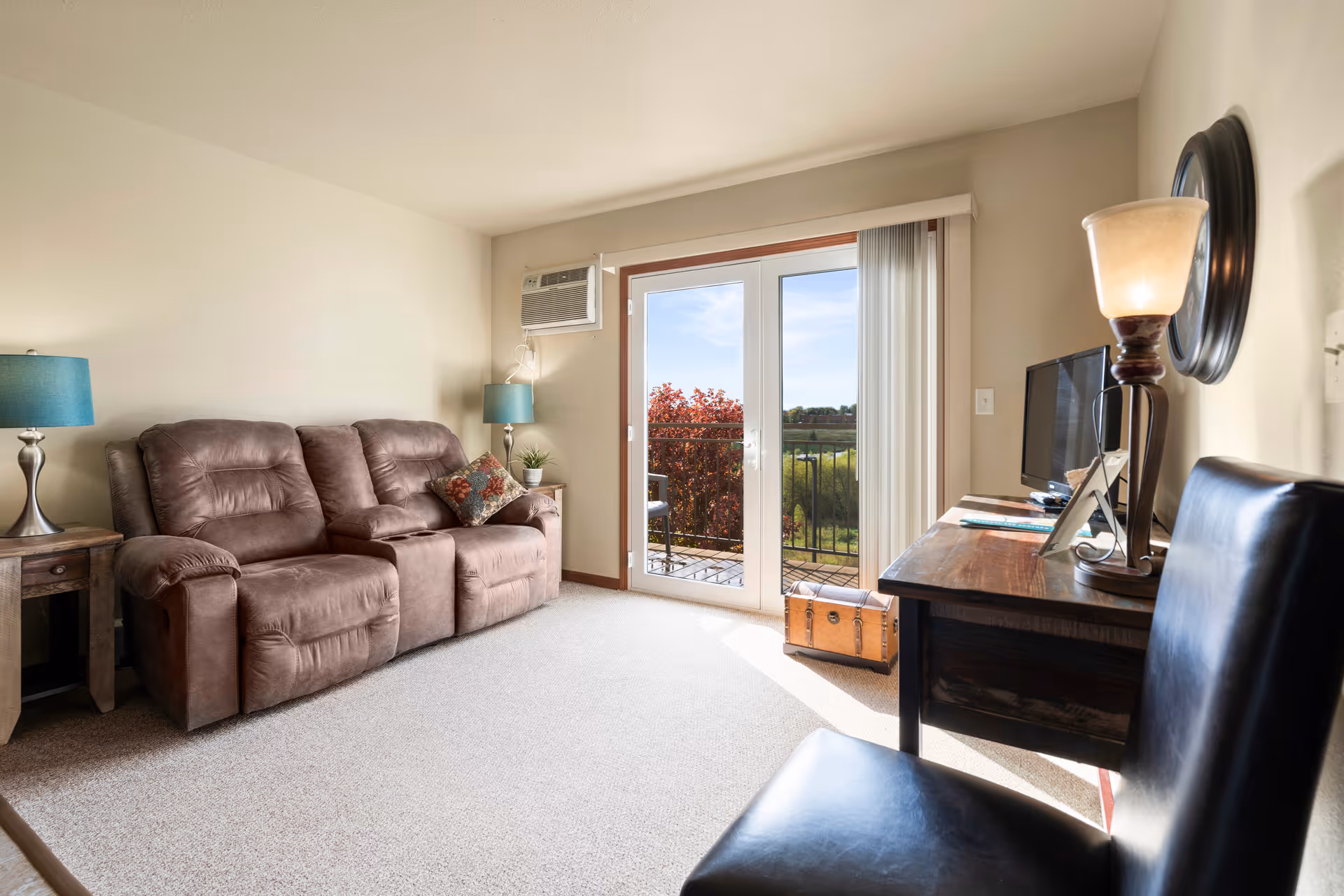 A cozy living room with a brown reclining sofa, two side tables with blue lamps, a wooden desk with a TV and a lamp, a black chair, and a glass door leading to a balcony with a view of greenery and a clear sky.