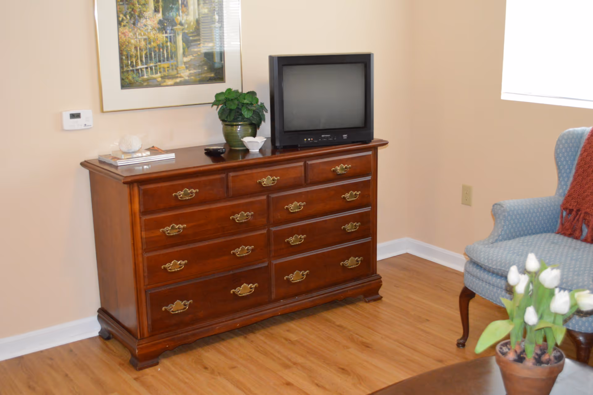 A wooden dresser with multiple drawers and brass handles stands against a beige wall. On top of the dresser is a small, old-style television, a green potted plant, a small white decorative bowl, and some books. To the right, there is a blue upholstered armchair with a red throw draped over one arm. A pot of white tulips is visible in the foreground on a wooden table. A framed painting hangs on the wall above the dresser, and a thermostat is mounted on the wall to the left.