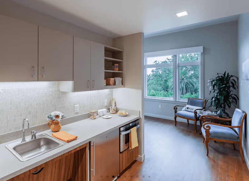 Small kitchenette with a sink, countertop and cabinets opening into a bright sitting area with two chairs by a large window.