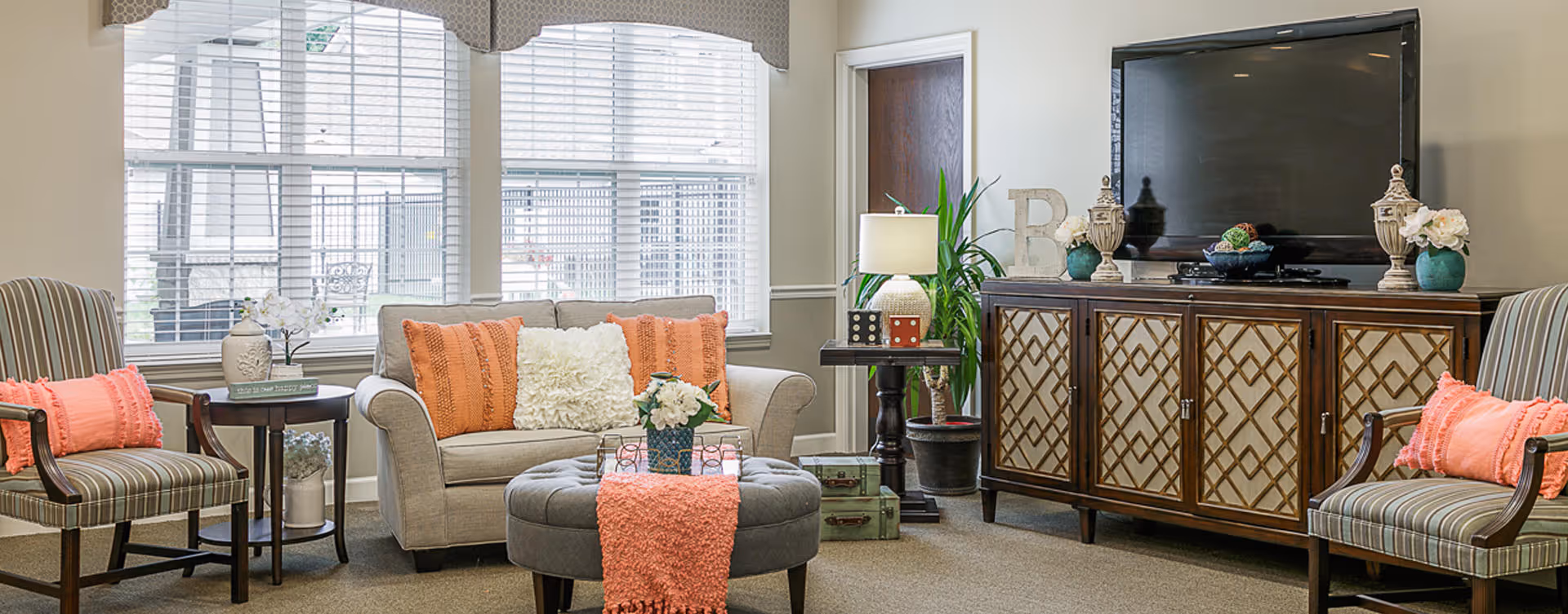 Cozy living room with a sofa, striped armchairs, an ottoman, TV on a wooden cabinet, and large windows.