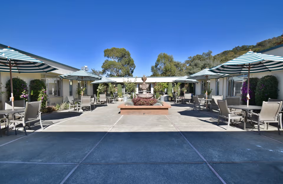Outdoor courtyard area with multiple tables and chairs under green and white striped umbrellas, surrounded by a single-story building with windows and greenery, and a central water fountain with plants. Trees and hills are visible in the background under a clear blue sky.