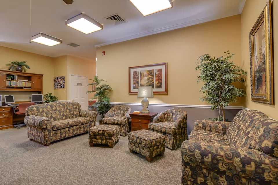 A comfortable senior living common room with patterned sofas, armchairs, ottomans, a lamp, framed artwork, plants, and a computer desk in the background.