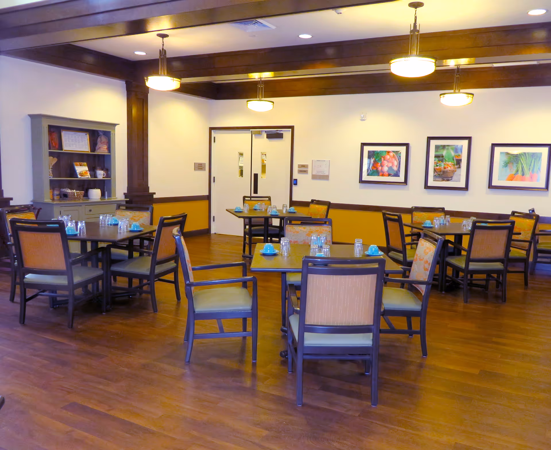 A dining room in Lakewood Memory Care facility with several tables and chairs arranged neatly. Each table has glasses and blue cups placed on it. The room has wooden flooring, warm lighting from ceiling fixtures, and framed pictures of fruits and vegetables on the wall. There is a cabinet with shelves holding various items in the corner.
