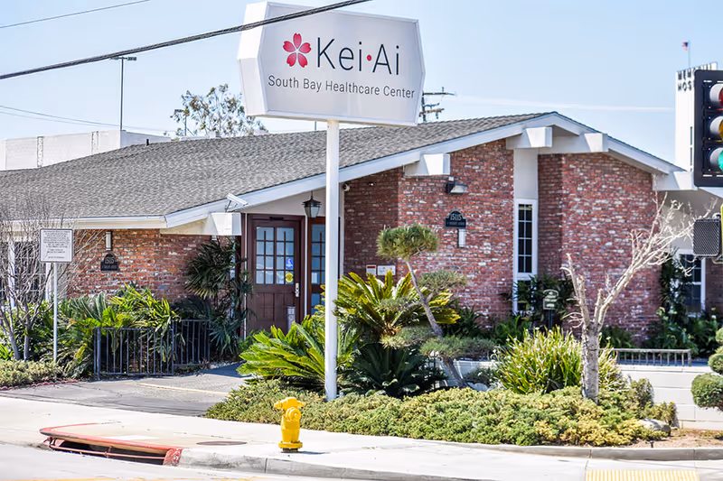 Exterior view of Kei-Ai South Bay Healthcare Center, a single-story brick building with a shingled roof, surrounded by green shrubs and plants. A large sign with the facility's name is mounted on a white pole near the sidewalk. A yellow fire hydrant is visible on the curb, and a traffic light is partially visible on the right side of the image.