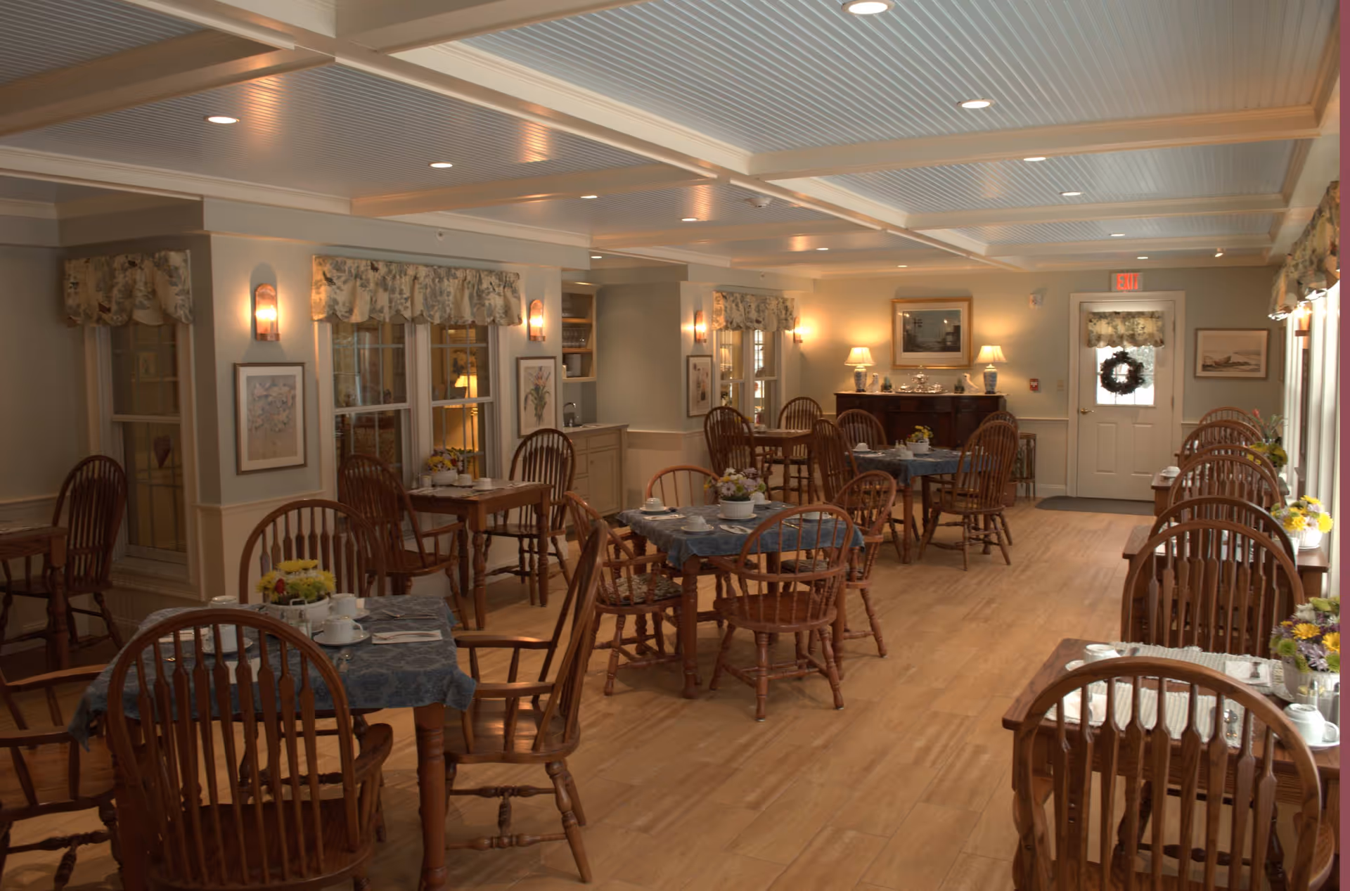 A cozy dining room in Decatur House with multiple wooden tables and chairs arranged neatly. Each table is covered with a blue tablecloth and set with white cups, saucers, and utensils. The room has soft lighting from wall sconces and table lamps, floral curtains on the windows, and framed artwork on the walls. The floor is wooden, and there is a door with a wreath at the far end of the room.