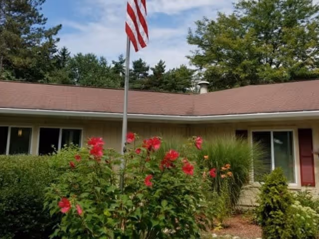 Single-story building with a brown roof and beige walls, surrounded by green bushes and red flowers in the foreground. An American flag is mounted on a pole in front of the building, with trees and a partly cloudy sky in the background.