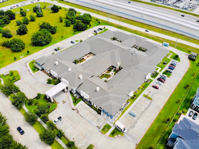 Aerial view of Orchard Park at Southfork Assisted & Senior Living facility showing a large building with a gray roof surrounded by parking lots, green lawns, trees, and nearby highways.
