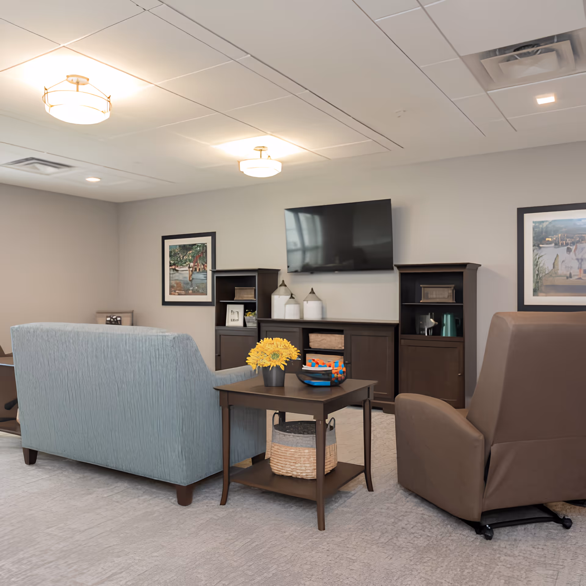 A senior living facility common area with a light blue sofa, a brown recliner chair, a wooden side table with a basket and a vase of yellow flowers, two dark wooden cabinets, a flat-screen TV mounted on the wall, and framed artwork on the walls.
